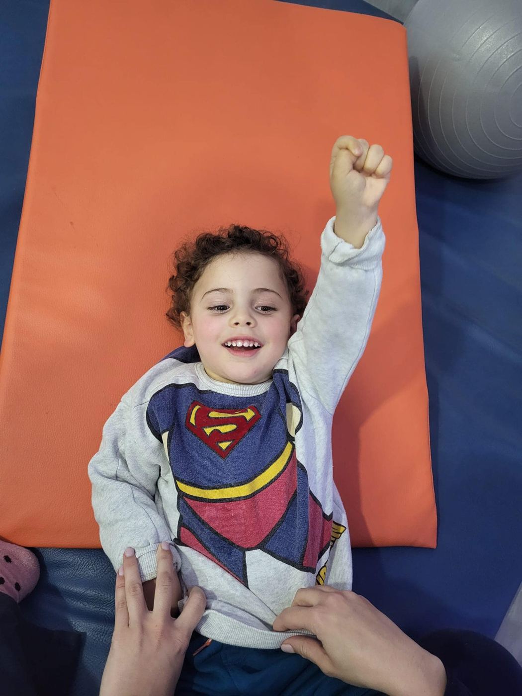 O Martim deitado num tapete laranja, de braço no ar, com uma camisola do Super-Homem