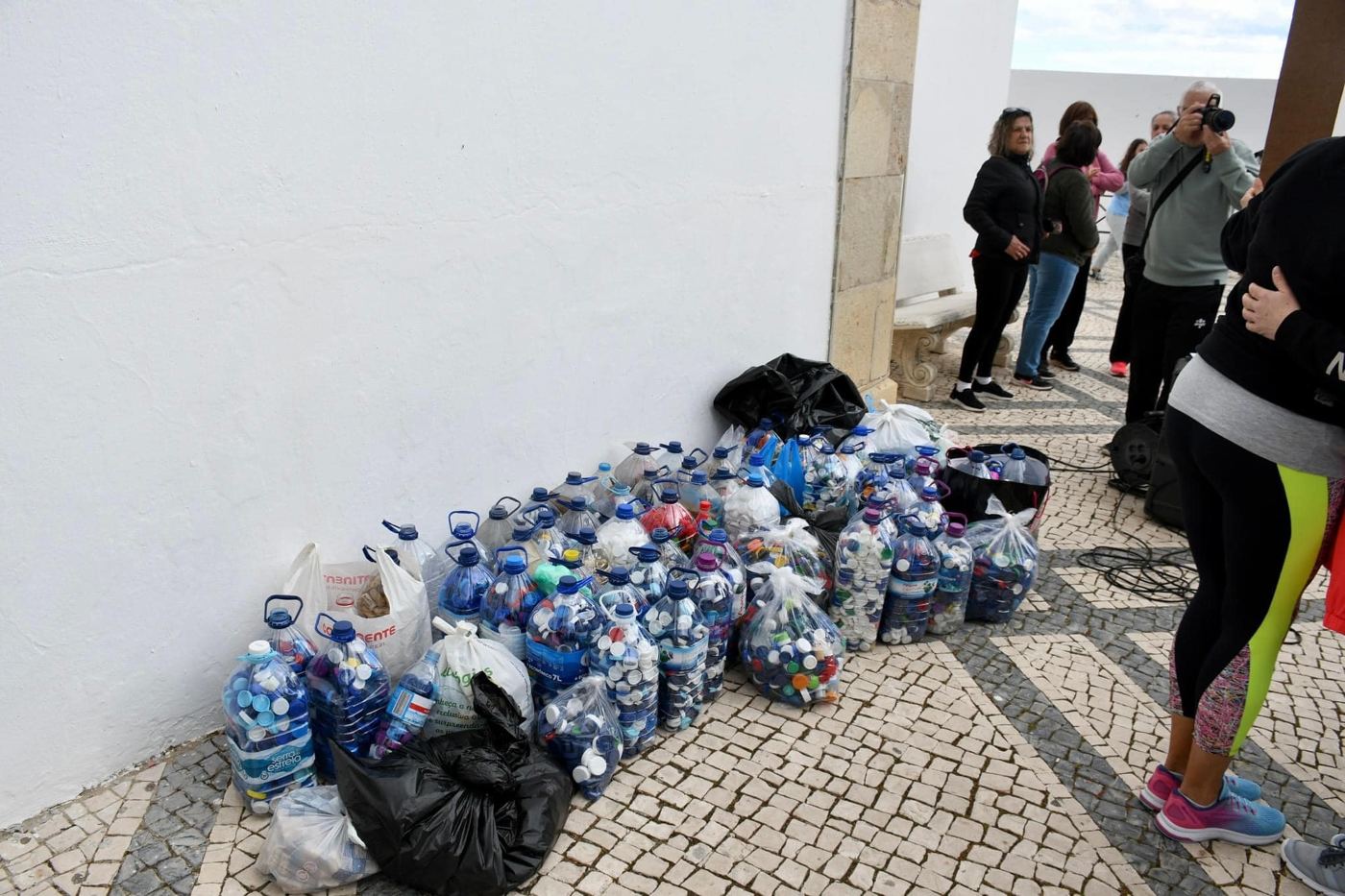 Sacos de tampas junto a uma parede, com pessoas a passar